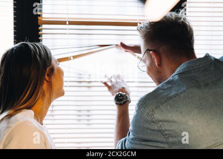 Young cheerful couple looks out the window through the blinds Stock Photo