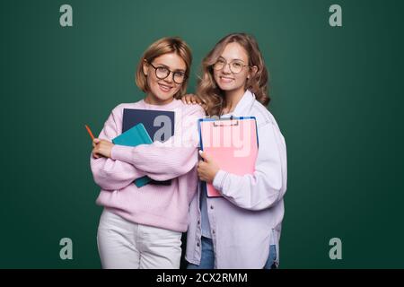 Two students posing with some folders and smile at camera wearing eyeglasses on a green studio wall Stock Photo