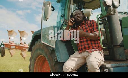 Young african farmer speaking on the smartphone while leaning on the green tractor. High quality photo Stock Photo
