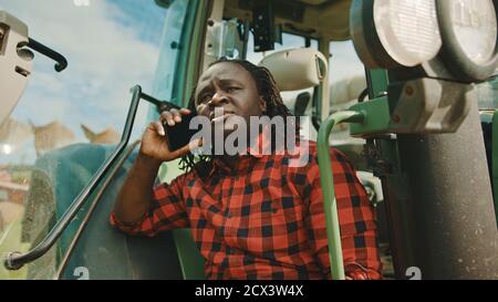Young african farmer speaking on the smartphone while leaning on the green tractor. High quality photo Stock Photo