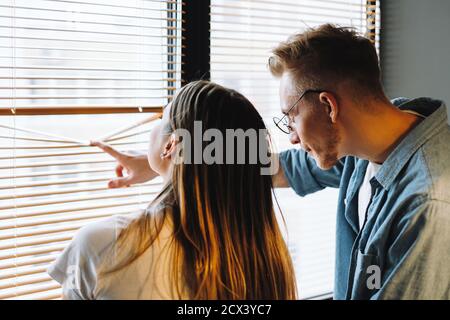 Young cheerful couple looks out the window through the blinds Stock Photo