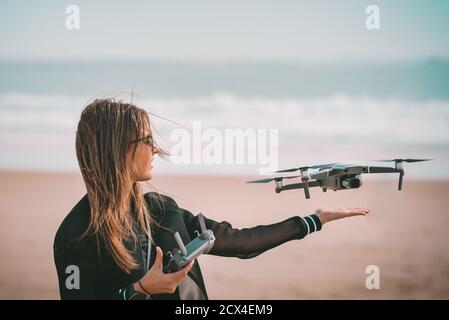 Young modern lady pilot in black jacket lands drone on her hand with remote controller near the ocean. Outdoors hobby. Stock Photo