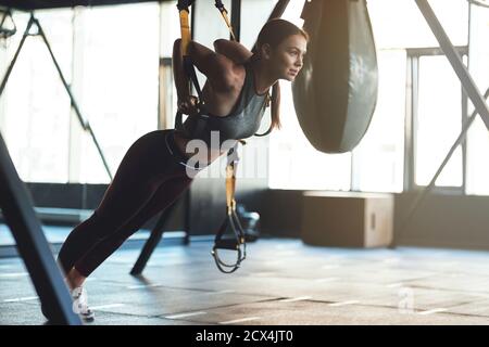 Young beautiful athletic woman wearing sports clothes exercising with fitness trx straps, doing push ups while training at gym. Sport, workout, wellness and healthy lifestyle Stock Photo