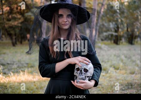 Young beautiful cute woman in dark dress and witch's hat holds a skull in her hands. Halloween party costume. Forest, park with autumn trees