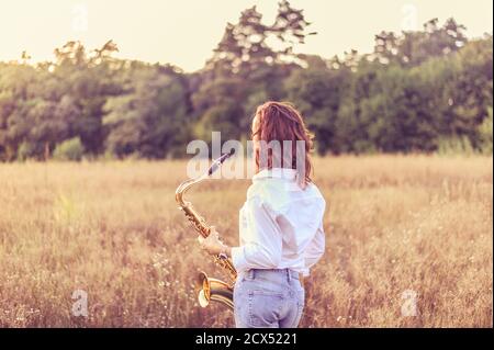 Young red-haired woman in a white shirt with a tenor saxophone in her hands stands in an autumn field and looks at the sunset Stock Photo