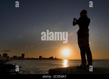 Western tourist standing on a pillar and photographing the Malecon at sunset, Havana, Cuba. MODEL RELEASED SUBJECT Stock Photo