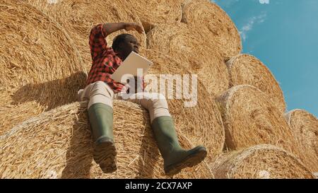 African black farmer sitting on the haystack and holding laptop. Making shade with hand over the face while looking at the distance. High quality photo Stock Photo