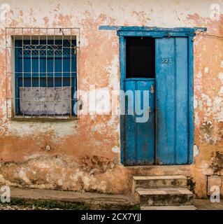 Trinidaad, Cuba Nov 26, 2017 - Half-open blue door plus window with grate on tattered yellow wall with stone steps Stock Photo