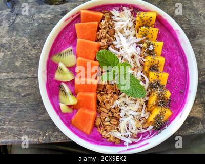 Purple dragon fruit smoothie bowl topped with fresh papaya, mango, kiwi, shredded coconut, chia seeds, granola and a sprig of mint with a wooden table Stock Photo