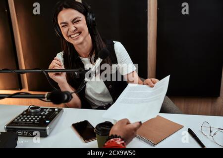 Portait of happy female radio host smiling, receiving a script paper from her male colleague while moderating a live show in studio Stock Photo