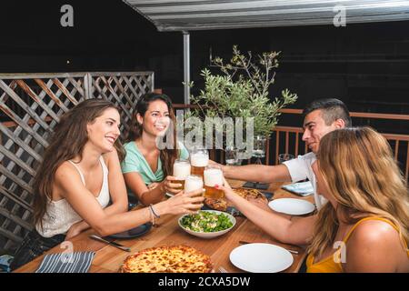 Friends cheering with beer glasses sitting around a table - Group of multiracial people toasting and clinking drinks at each others Stock Photo