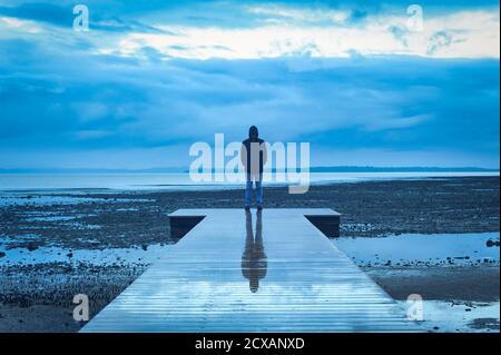 A blue toned,  fine art image of a single male standing on a pier at sunset with a brewing thunderstorm out to sea at Cleveland Point, Australia Stock Photo