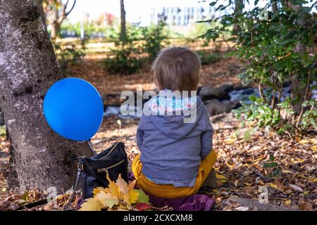 Photo of a little lonely boy in a grey jacket and orange pants sitting on the ground next to a park creek. Blue balloon tied to a bag next to him. Stock Photo