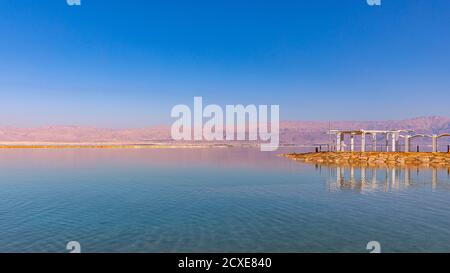 Scenic view at calm waters of  Dead Sea, Israel.  Sea beach with structures reflecting in calm water against blue sky and mountains in the background Stock Photo