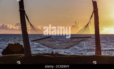 Empty hammock between two palm trees against scenic sunset over Pacific ocean. Calm ocean and clouds above the horizon, Big Island, Hawaii Stock Photo