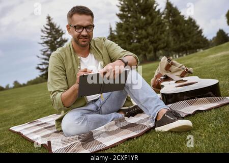 Writing a song. Young creative man wearing eyeglasses sitting on a green grass in park and making some notes, acoustic guitar lying near him Stock Photo