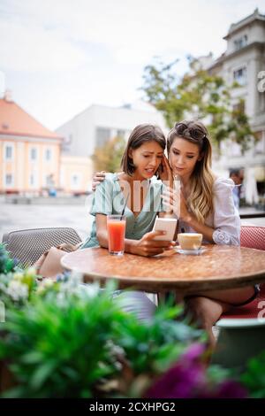Depressed young woman holding mobile phone while being consoled by her friend Stock Photo