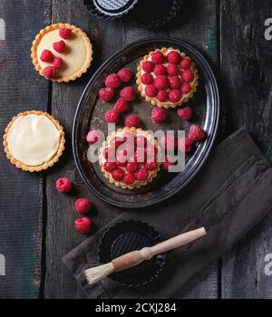 Unfinished and ready to eat tartlets with custard and fresh ripe raspberries, served on vintage metal tray with baking forms and brush for glazing ove Stock Photo