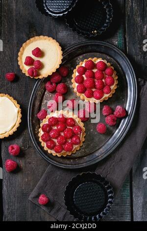 Unfinished and ready to eat tartlets with custard and fresh ripe raspberries, served on vintage metal tray with baking forms and textile napkin over o Stock Photo
