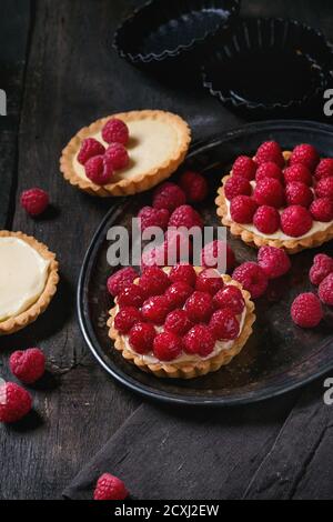 Unfinished and ready to eat tartlets with custard and fresh ripe raspberries, served on vintage metal tray with baking forms and textile napkin over o Stock Photo