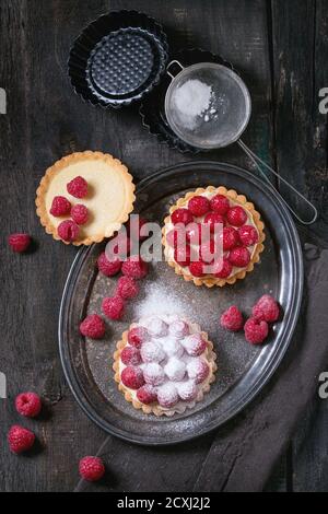 Unfinished and ready to eat tartlets with custard, sugar powder and fresh raspberries, served on vintage metal tray with baking forms and sieve over o Stock Photo