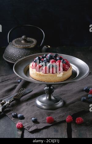 Homemade tart with custard, fresh raspberries and blueberries, served on vintage metal plate with iron teapot on textile napkin over old wooden table. Stock Photo