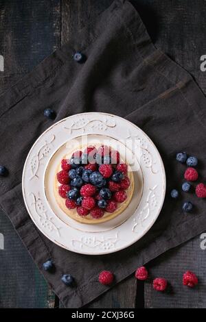 Homemade tart with custard, fresh raspberries and blueberries, served on white vintage plate on textile napkin over old wooden table. Dark rustic styl Stock Photo