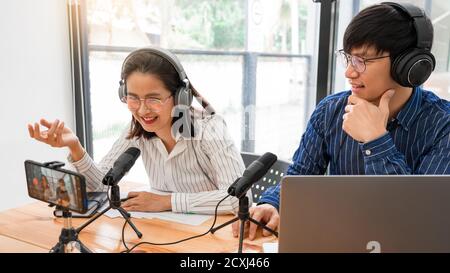 Asian Man and woman podcasters in headphones recording content with colleague talking to microphone and camera in broadcast studio together, communica Stock Photo