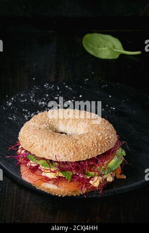 Bagel with salted salmon, spinach, beetroot sprouts, avocado and scrambled egg on wooden chopping board over old wood black background. Stock Photo