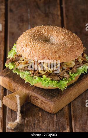 Bagel with stew beef, fresh salad and fried onion on small wooden chopping board over wooden background. Stock Photo