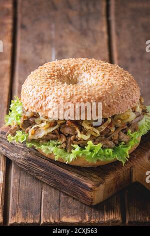 Bagel with stew beef, fresh salad and fried onion on small wooden chopping board over wooden background. Stock Photo