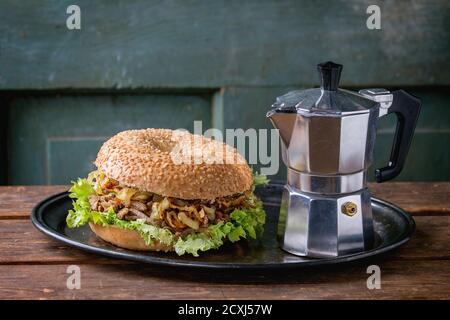 Bagel with stew beef, fresh salad and fried onion served on vintage metal tray with coffee pot over wooden table. Stock Photo