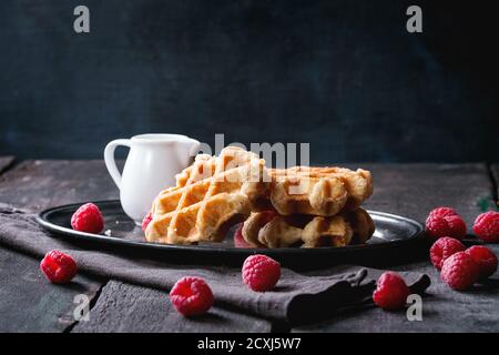 Belgian waffles with raspberries, served with jug of milk on vintage metal tray with textile napkin over old wooden table. Dark rustic style. Stock Photo