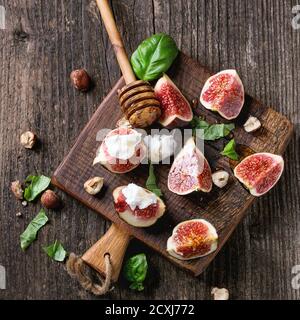 Fresh sliced figs with ricotta cheese, basil leaves, hazelnut and honey from honey dipper on small wood cutting board over dark wooden background. Top Stock Photo