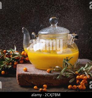 Hot berry sea buckthorn tea drink with apples in glass teapot on clay board, served with glass cup, saucer, fresh berries on branch over old wooden ba Stock Photo