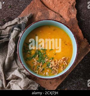 Blue ceramic bowl of carrot soup, served with onion chives and kitchen towel on clay board over old rusty iron background. Dark rustic style. Top view Stock Photo