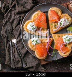 Wafers as heart shape with salted salmon, red onion, chive, lime and ricotta cheese on metal plate with vintage cutlery and sackcloth rag over dark ru Stock Photo