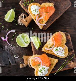 Wafers as heart shape with salted salmon, red onion, chive, lime and ricotta cheese on small wooden cutting boards over dark wood background. Top view Stock Photo