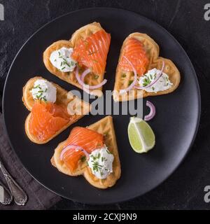 Wafers as heart shape with salted salmon, onion, chive, lime and ricotta cheese on black plate with vintage cutlery and textile napkin over black ston Stock Photo