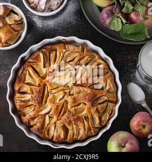 Homemade big and small apple cake pie in white ceramic forms with fresh apples with leaves, sugar powder, and tin can of sugar over dark wooden backgr Stock Photo