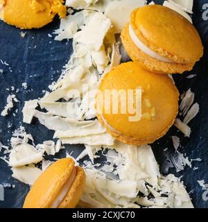 Whole and broken orange lemon homemade macaroons with chopped white chocolate and citrus sugar and zest on dark glass board over black textural backgr Stock Photo