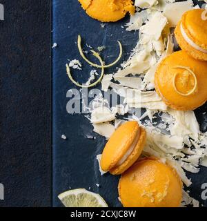 Whole and broken orange lemon homemade macaroons with chopped white chocolate and citrus sugar and zest on dark glass board over black textural backgr Stock Photo