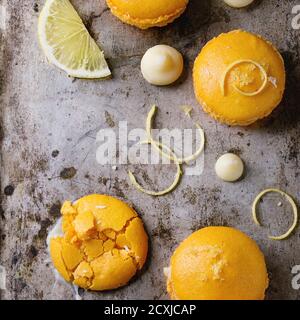Whole and broken orange lemon homemade macaroons with chopped white chocolate and citrus sugar and zest over old metal textural background. Overhead v Stock Photo