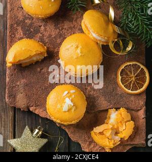 Whole and broken orange lemon homemade macaroons with white chocolate and citrus sugar and zest on old clay board over dark wooden background with Chr Stock Photo
