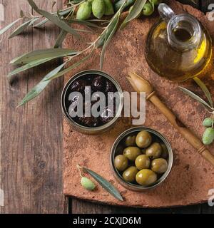 Green and black olives in tin cans, young olives branch and bottle of olive oil on clay board over old wood background. Overhead view with space for t Stock Photo