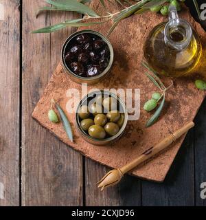 Green and black olives in tin cans, young olives branch and bottle of olive oil on clay board over old wood background. Overhead view with space for t Stock Photo