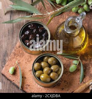 Green and black olives in tin cans, young olives branch and bottle of olive oil on clay board over old wood background. Overhead view with space for t Stock Photo