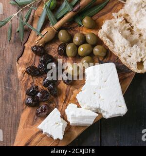 Green and black olives with loaf of fresh bread, feta cheese and young olives branch on olive wood chopping board over old wood background. Overhead v Stock Photo