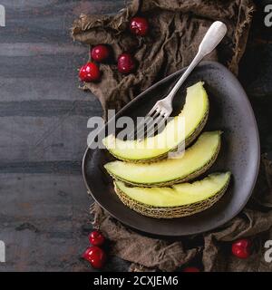 Brown ceramic plate with sliced melon, served with cherry berries and vintage fork on wet sackcloth rag over old dark textured iron background. Top vi Stock Photo