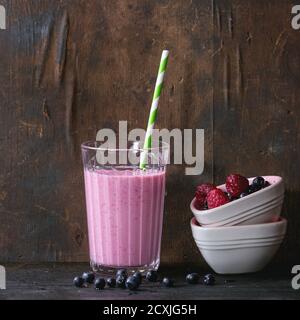 Glass of homemade raspberry smoothie, served with cocktail tube and bowl of fresh berries over old dark wood background. Healthy breakfast theme. With